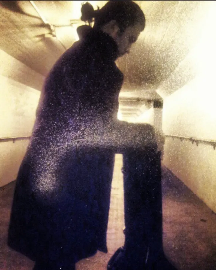 Grainy instant-photo style picture of a man in silhouette with his hair in a bun, leaning on an upright instrument case in a tunnel